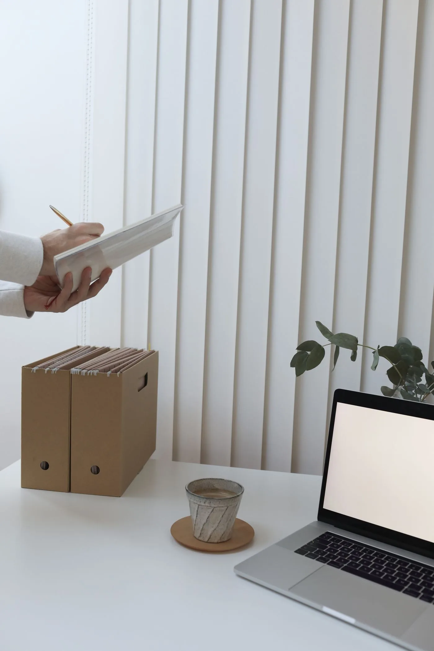 白い机に置かれた書類ボックスとノートパソコン、冊子を手にした人の腕